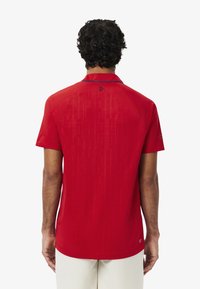 Punainen lyhythihainen polo-paita, jossa on teksturoitu kuvio, terävä kaulus ja kontrastiväriset laivastonsiniset yksityiskohdat kauluksessa ja hihoissa.
