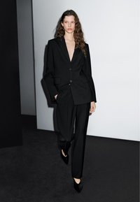 Zwart op maat gemaakt pak met een oversized blazer met twee knopen, wijde broekspijpen en puntige zwarte hakken. Soepele textuur en minimalistisch ontwerp.