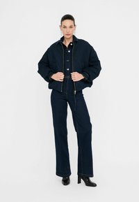 Giacca di jeans scura trapuntata con chiusura a zip e dettagli con bottoni, abbinata a pantaloni di jeans scuri a vita alta e stivaletti neri con tacco.