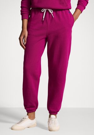Persoon die magentakleurige joggingsbroek draagt met witte aantrekkoord en beige sneakers, staand met één hand in de zak tegen een witte achtergrond.