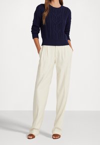 Pull marin en maille avec motif torsadé, associé à un pantalon plissé crème ; les deux portés avec des sandales marron à bout ouvert. Textures lisses, coupe décontractée.