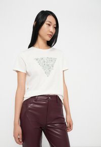 Camiseta blanca con un detalle de bordado floral plateado triangular en el centro, combinada con pantalones de cuero sintético color burdeos.