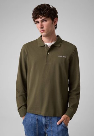 Jeune homme aux cheveux bouclés portant un polo à manches longues vert olive avec le logo "GUESS JEANS" et un jean bleu, les mains dans les poches.
