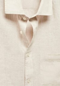Jasnobeżowa lniana koszula z kołnierzykiem zapinanym na guziki, z małą kieszonką na piersi i subtelną fakturą. Pięć guzików z przodu.