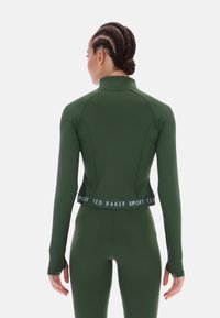 Zöld sportos felső magas nyakrésszel és hosszú ujjakkal, testhez álló fazonban és márkás derékpánttal. Simább, rugalmas anyagból.