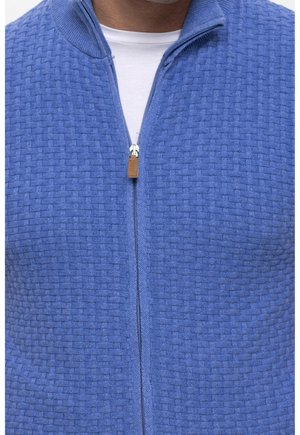 Mężczyzna ubrany w niebieski sweter zapinany na zamek o fakturze, na biały t-shirt z okrągłym dekoltem, z zamkiem zaciągniętym przy klatce piersiowej.