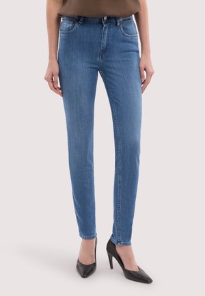 Kvinde iført smalle blå jeans og sorte spidse højhælede sko, stående mod en lys ensfarvet baggrund.