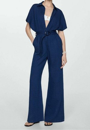 Kobieta ubrana w granatowy kombinezon z szerokimi nogawkami i paskiem, z krótkimi rękawami i białą bluzką pod spodem, stojąca z rękami w kieszeniach.