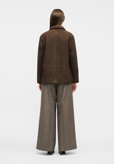 Γυναίκα που στέκεται με θέα προς τα πίσω, φοράει ένα σκούρο καφέ σακούλι από σουέτ και φαρδιά γκρι παντελόνια σε απλό λευκό φόντο.