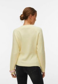 Femme aux cheveux bruns attachés en chignon, portant un pull jaune pâle et un jean noir, debout de dos devant un fond blanc.