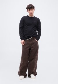 Jeune homme debout, les mains dans les poches, portant une chemise noire à manches longues, un pantalon large brun et des baskets blanches sur fond blanc.