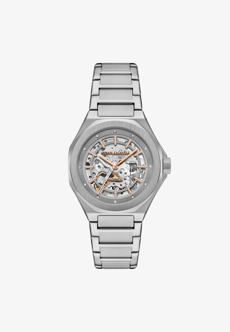 Ασημί ρολόι από ανοξείδωτο ατσάλι με διάφανο καντράν, με πορτοκαλί λεπτομέρειες και βραχιόλι από ματ μέταλλο. Αυτόματη κίνηση ορατή μέσω της πρόσοψης.