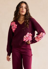 Maglione a maglia bordeaux con scollo a V, decorato con disegni di rose in paillettes rosa sulle maniche. Abbinato a pantaloni bordeaux coordinati.
