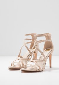 Beiga högklackade sandaler med remmar och en slank design. Slät mockaskin, justerbara ankelsremmar och öppen tå-stil med minimal hårdvara.