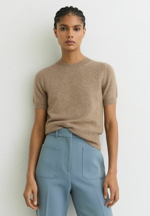 Kvinde med flettet hår iført en beige kortærmet sweater, der er stoppet ned i højtaljede lyseblå bukser med lommer foran.