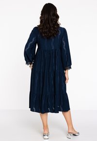 Marineblaues, schichtenförmiges Kleid mit langen Puffärmeln, das spitzenähnliche Ausschnitte und einen fließenden Rock hat, kombiniert mit silbernen Mules.