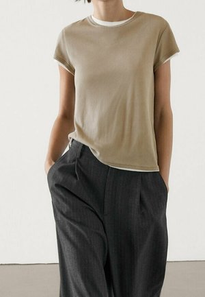 Person wearing a beige short-sleeve shirt layered over white, paired with loose dark gray pinstripe pants, hands in pockets.