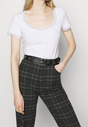 Frau trägt ein weißes, kurzärmeliges Oberteil mit Spitzenbesatz, das in hoch taillierte schwarz-grau karierten Hosen mit schwarzem Gürtel gesteckt ist.