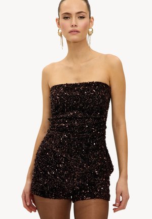 Femme portant une combinaison courte bustier à sequins foncés avec des boucles d'oreilles dorées remarquables, posant devant un fond clair uni.