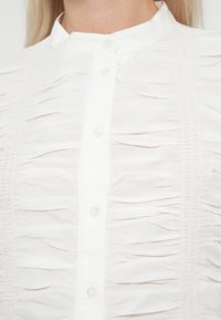 Hvid bluse med flæset stofpaneler og knapper foran, båret af en person med blondt hår.