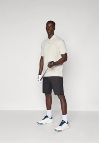 Krémové polo tričko a černé šortky, které jsou doplněny bílými a námořnickými golfovými botami. Osoba drží golfovou hůl a má na sobě golfovou rukavici.