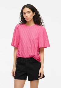 Blusa rosa con maniche corte a volant; tessuto testurizzato con motivo a diamante; indossata con pantaloni corti in denim neri.