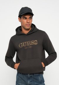 Felpa grigia scura con tasca frontale, con il logo dorato "GUESS" e il testo "EST. 1981". Abbinata a un berretto nero. Vestibilità casual.
