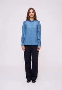 Blusa in satin azzurro chiaro con colletto, chiusura a bottoni e maniche lunghe a sbuffo, abbinata a pantaloni larghi scuri e scarpe marroni.
