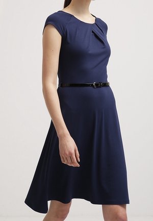Kobieta w granatowej sukience do kolan z krótkimi rękawkami i cienkim czarnym paskiem w talii, stojąca na jednolitym tle.