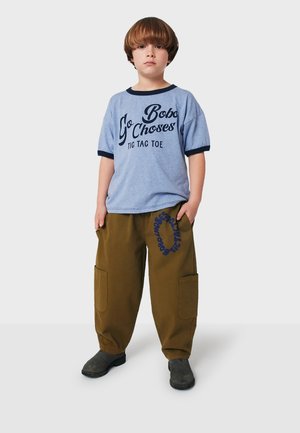 Light blue t-shirt with dark blue trim featuring printed text; loose brown cargo pants with side pockets and a blue circular logo. Black shoes.