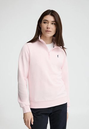 Młoda kobieta w jasno różowej bluzie z zamkiem do połowy i ciemnych spodniach stoi na tle jednolitego jasnego tła.