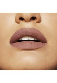 Rouge à lèvres mauve mat sur des lèvres pulpeuses, avec des détails de texture visibles. Le teint est chaud. Les dents sont partiellement visibles. Fini lisse.