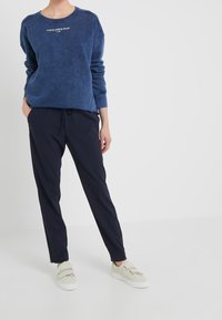 Felpa blu con scollatura rotonda e testo bianco, abbinata a pantaloni blu navy e scarpe beige. Il tessuto appare morbido con una vestibilità rilassata.