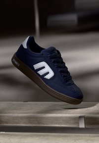 Μαύρο αθλητικό παπούτσι με λευκό λογότυπο και λεπτομέρειες. Διαθέτει υφασμάτινη δερμάτινη επάνω επιφάνεια, σχέδιο με κορδόνια και καφέ λαστιχένια σόλα.