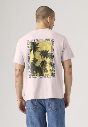 Hombre vestido con camiseta rosa clara con gráfico de palmera en negro y amarillo y texto "Levi's Coastal State of Mind" en la espalda, combinado con vaqueros azules.