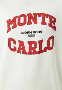 Krém színű póló, amelyen merész piros felirat található, amely így szól: "MONTE CARLO" és "BJÖRN BORG 1956" feketével, sima textúrával.