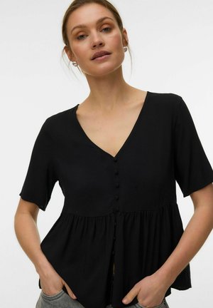 Kobieta o jasnej karnacji i jasnobrązowych włosach nosi czarną bluzkę z dekoltem w serek, zapinaną na guziki, z krótkimi rękawami i luźno marszczonym pasem, ręce w kieszeniach.