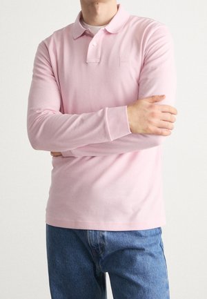 Mann trägt ein langärmliges rosa Poloshirt mit Kragen, steht mit verschränkten Armen, kombiniert mit blauen Denim-Jeans.