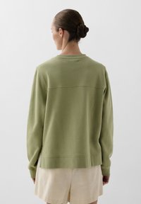 Grøn, langærmet sweatshirt i struktureret materiale med rund halsudskæring og subtile sømdetaljer, parret med lyse beige shorts.