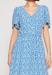 Donna che indossa un vestito blu con stampa floreale bianca, maniche corte, scollo a V, bottoni sul davanti e una cintura in vita.