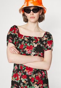 Vestido preto floral com rosas vermelhas e flores brancas, mangas curtas bufantes, decote quadrado. Combinado com um chapéu de balde em tie-dye laranja e óculos de sol pretos.