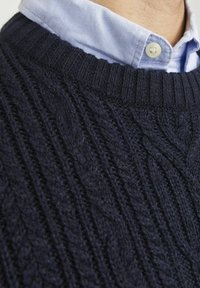 Navy cable-knit sweater with a ribbed neckline layered over a light blue collared shirt. Textured pattern adds depth.