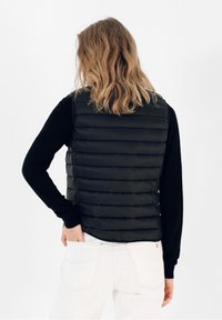 Sort puffervest med vandret syninger, båret over en langærmet sort skjorte. Modellen har bølget hår og hænderne i lommen på de hvide bukser.