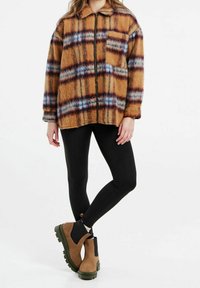 Veste-chemise marron à carreaux avec une fermeture éclair, arborant des motifs bleus et rouges. Associée à des leggings noirs et des bottines marron avec des semelles vertes.