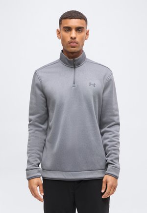 Νέος άνδρας με γκρι πουλόβερ Under Armour με φερμουάρ έως τη μέση και μαύρο παντελόνι, στέκεται μπροστά σε ένα απλό ανοιχτό φόντο.