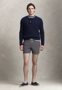 Modelul poartă un pulover tricotat bleumarin, pantaloni scurți gri și espadrile albastre. O cămașă cu dungi este parțial vizibilă. Ochelarii de soare sunt agățați de pulover.