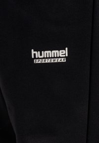 Zwarte sportbroek van zacht materiaal, met een wit logo "hummel SPORTSWEAR" aan de linkerkant. Eenvoudig ontwerp, geen patronen.