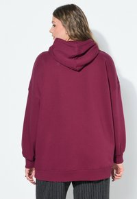 Mujer con cabello ondulado que lleva una sudadera holgada color marrón y pantalones negros a rayas, de espaldas contra un fondo blanco liso.