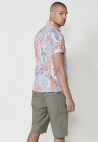 Camicia a maniche corte in tessuto leggero con un motivo di foglie grigie, arancioni e verdi; abbinata a pantaloni cargo color oliva con tasche laterali.