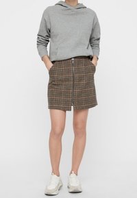 Sudadera gris combinada con una falda de cuadros marrón con cremallera frontal, que cuenta con bolsillos. La modelo lleva zapatillas de deporte de color claro. Diseño simple y casual.